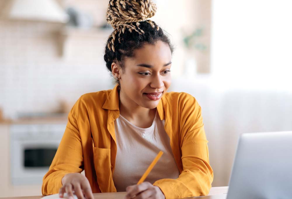 Photo of young woman at a desk and taking notes from her research on a laptop.